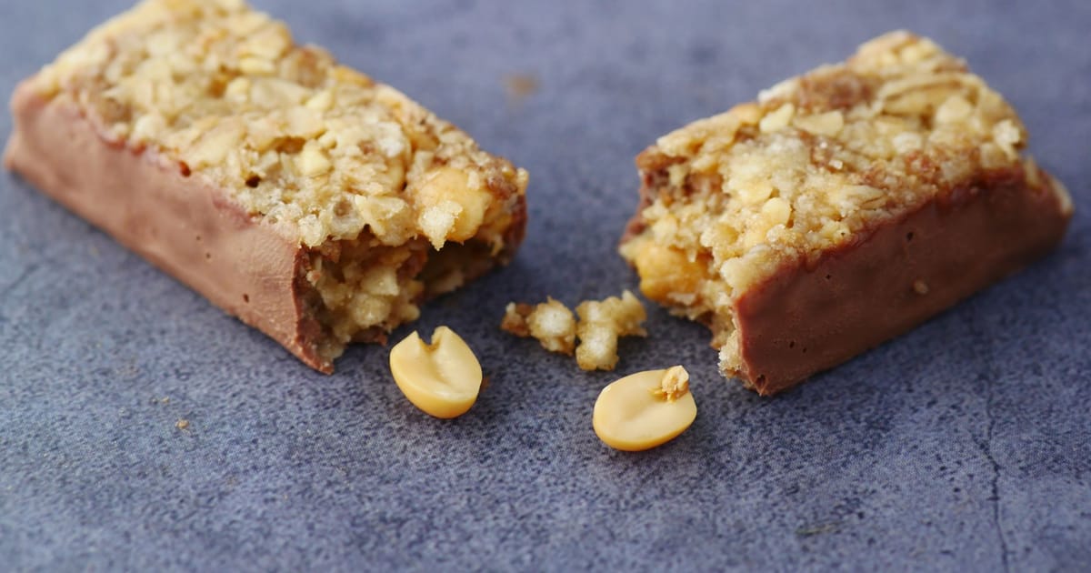 Barra de cereal coberta de chocolate partida ao meio, com amendoins soltos sobre fundo cinza