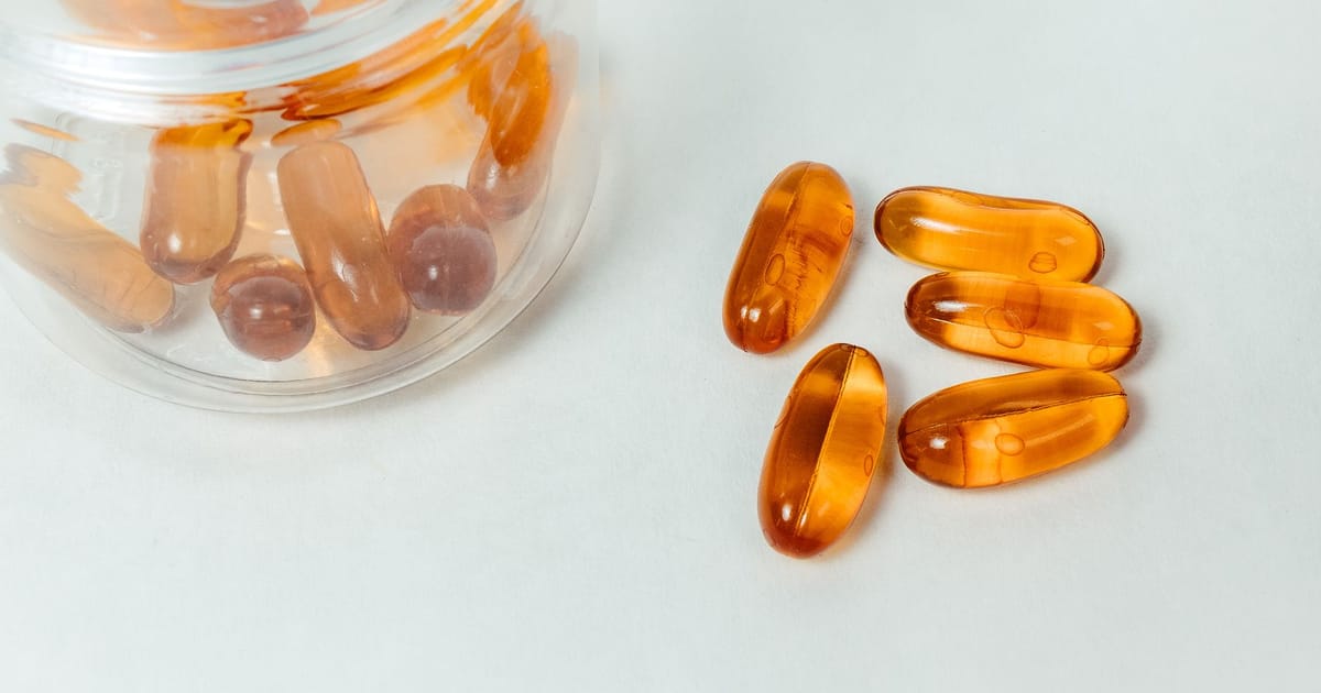 Cápsulas gel âmbar de suplemento saindo de um pote plástico sobre fundo claro