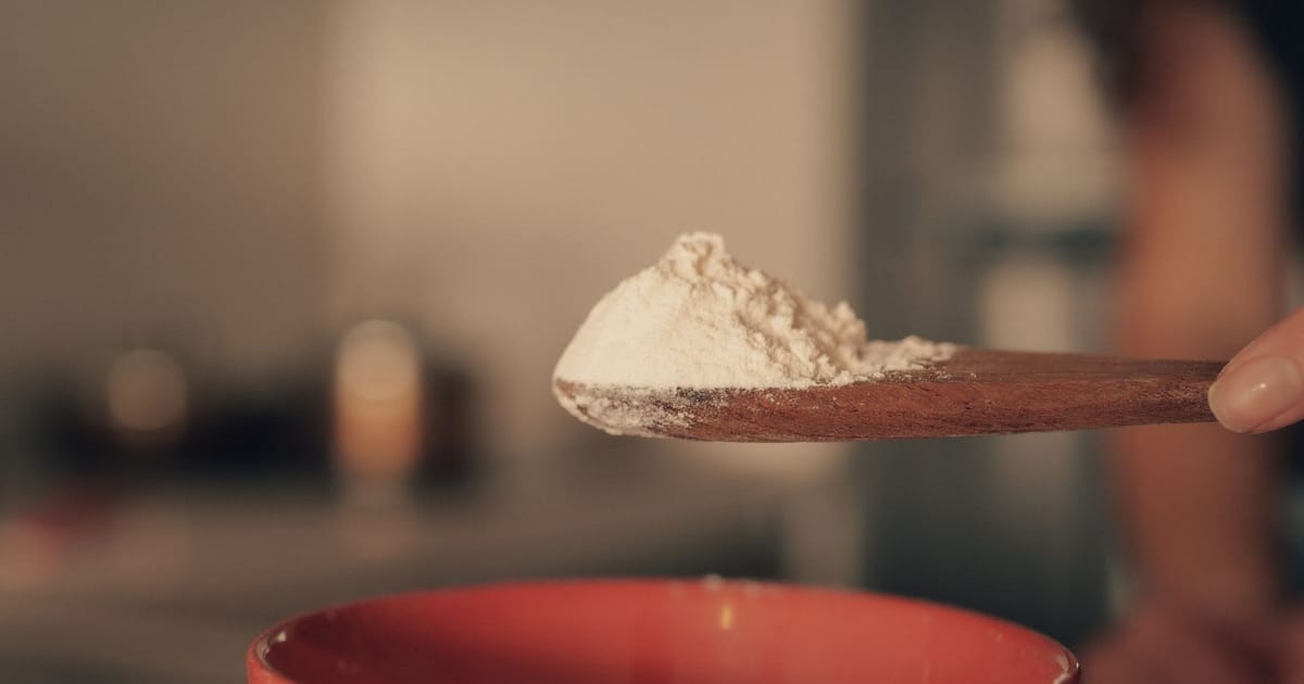 Colher de pau com pó branco de suplemento sendo levantada sobre uma tigela numa cozinha