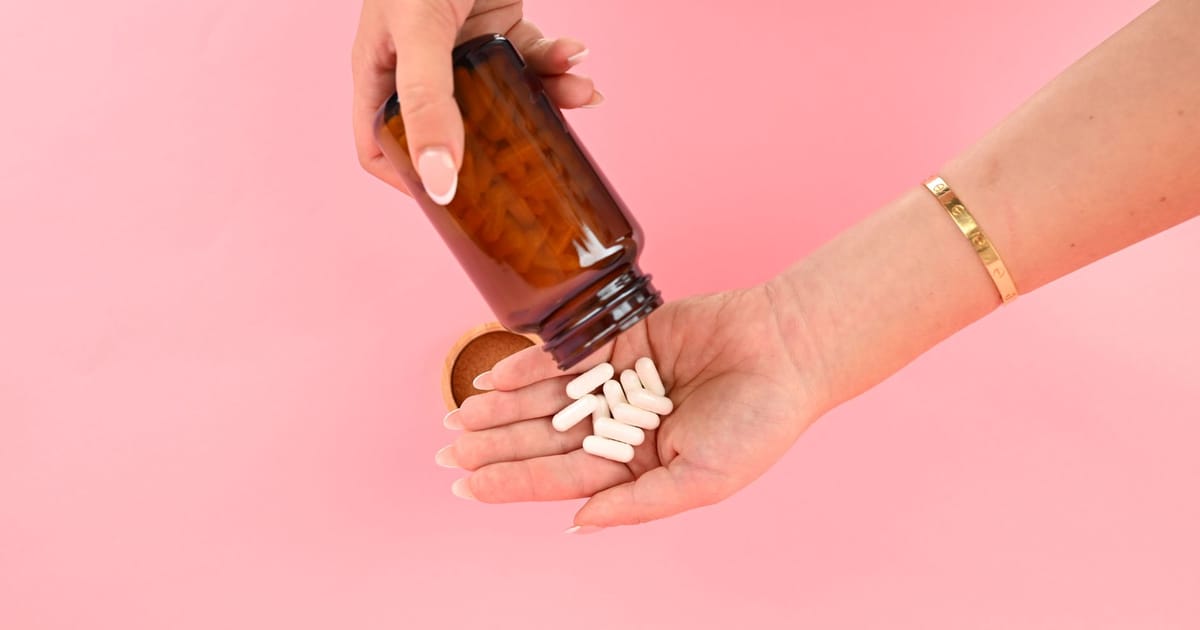 Mão despejando cápsulas brancas de suplemento de um pote âmbar sobre fundo rosa