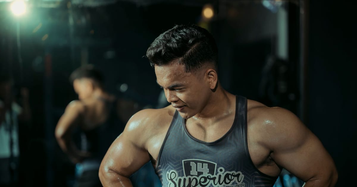 Homem musculoso de regata numa academia com iluminação escura, suado durante o treino