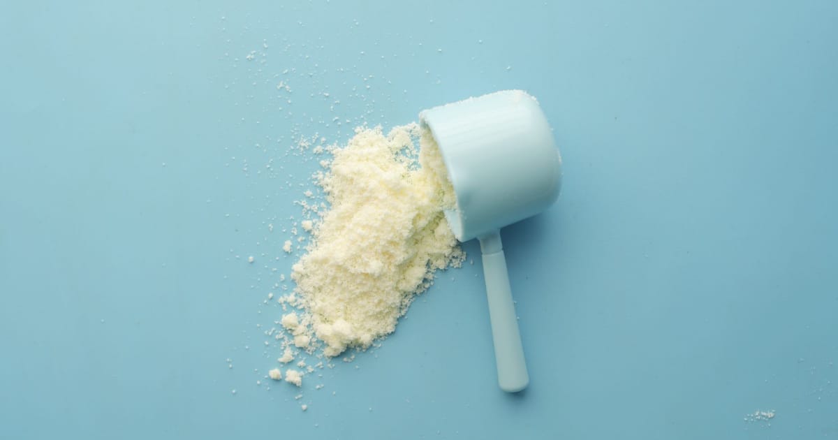 Colher dosadora branca com whey protein em pó derramado sobre um fundo azul claro