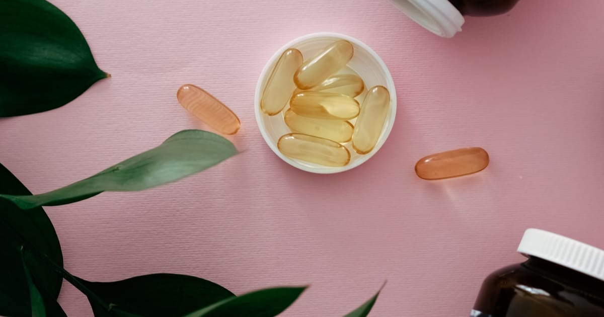 Cápsulas gel douradas numa tigela branca sobre fundo rosa, com folhas verdes ao lado